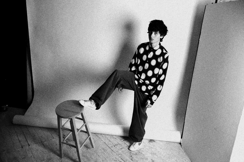 Marcel in a patterned shirt with one foot resting on a stool in the studio