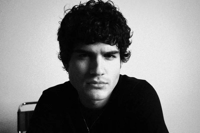 Pedro leaning toward the camera with curly hair and a dark shirt