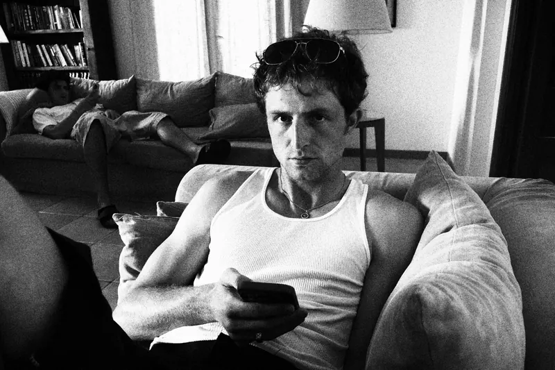 Max seated on a sofa in a white tank top, holding a phone